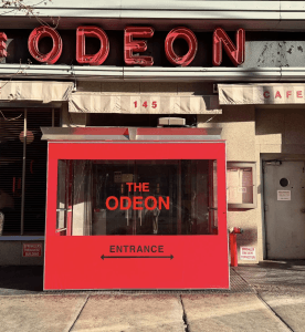 Bright red entrance booth with THE ODEON and ENTRANCE written on it stands outside a building, near Sylvias, with a large neon ODEON sign above and the address number 145 visible. Sunlight casts shadows on the sidewalk.